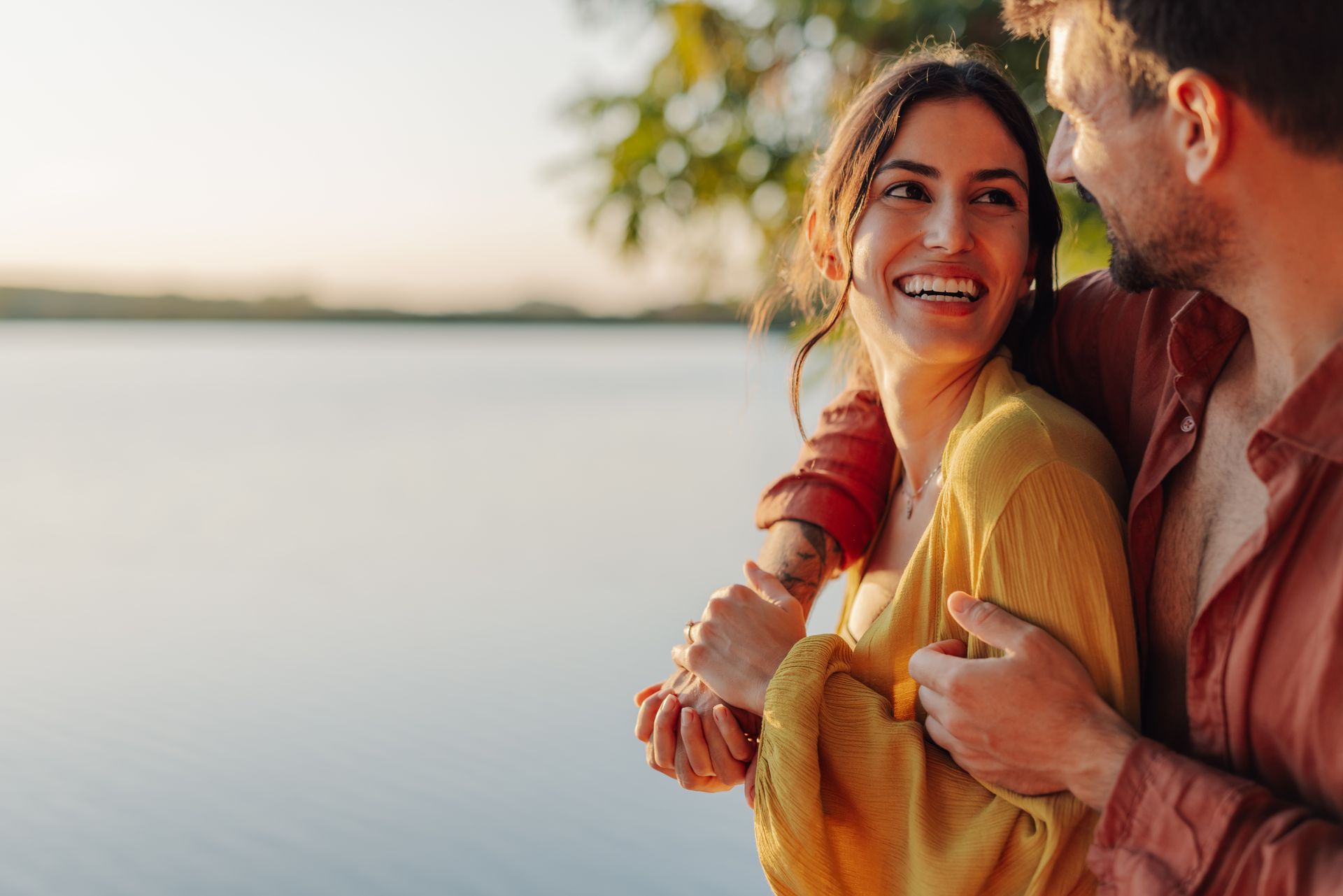 Couple embracing by water, woman laughing, warm colors.