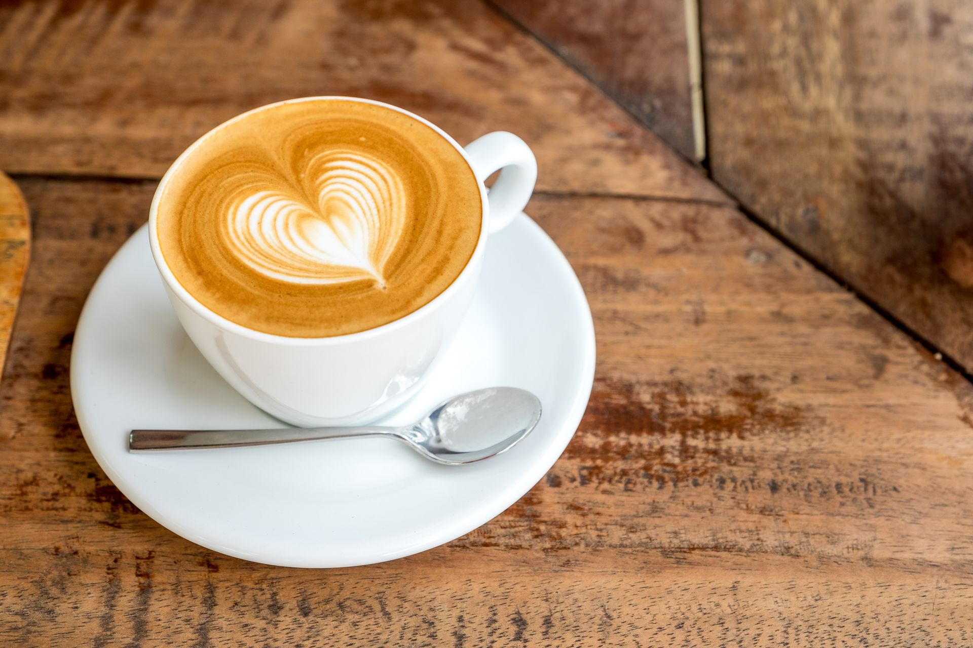 A cup of cappuccino with a heart shaped latte art on a saucer on a wooden table.