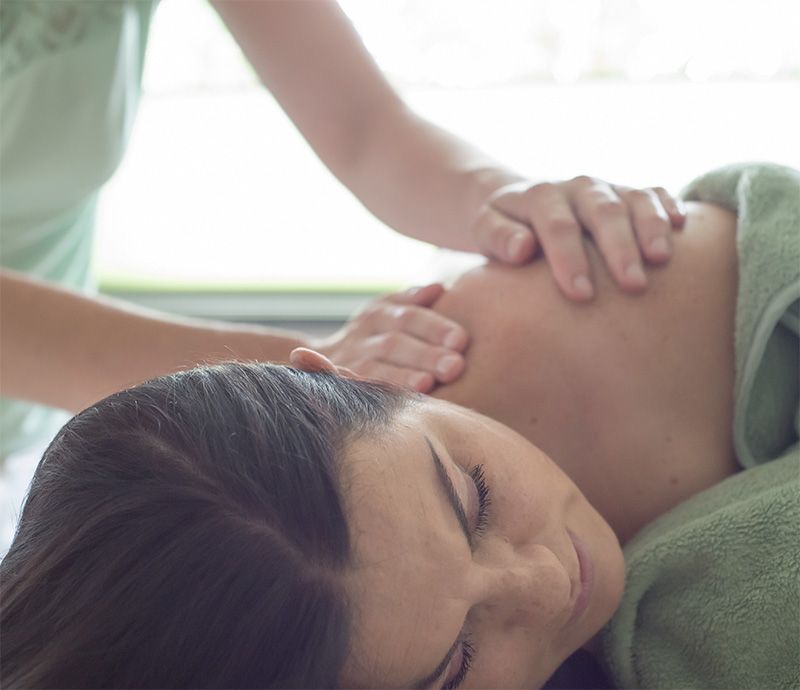 Vrouw krijgt een schoudermassage; ogen gesloten, ontspannen. Handen van de therapeut op de schouder; zachte groene handdoeken en kleding.