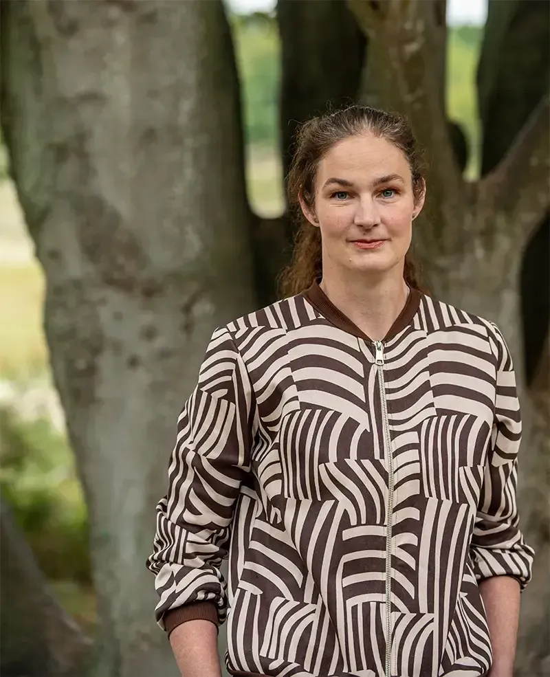 Annemarie in een bruin en crèmekleurig bomberjack, staand voor een boom.