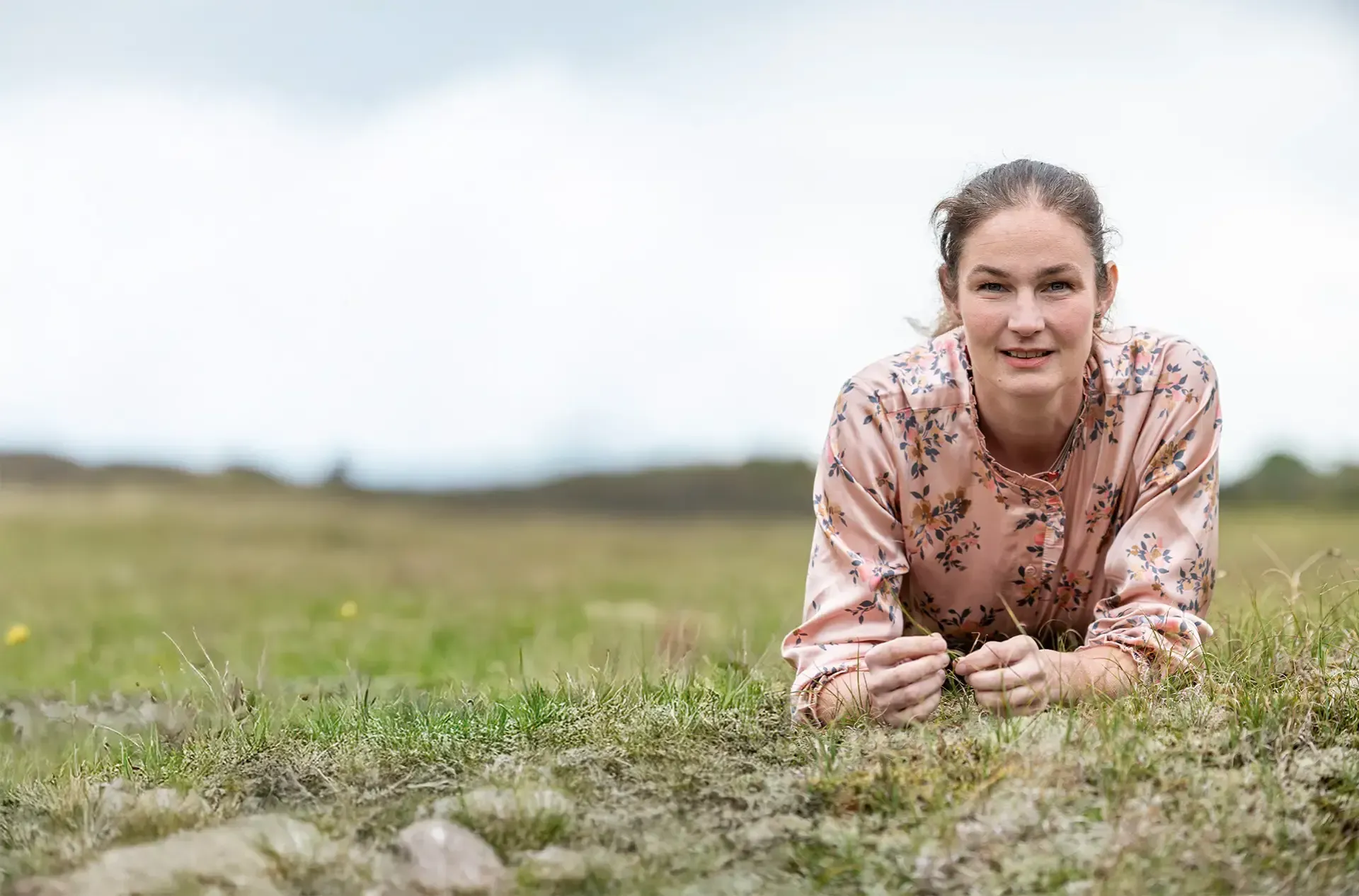 Annemarie in een roze bloemenblouse ligt lachend in het gras. Bewolkte lucht en een open veld op de achtergrond.