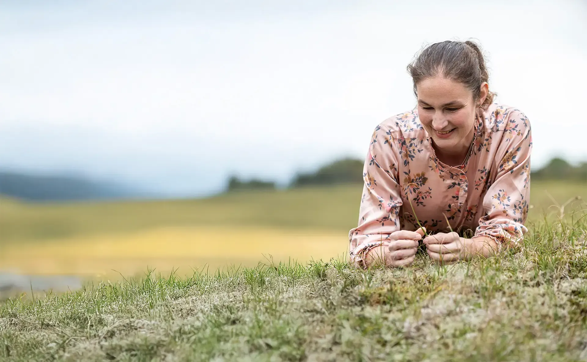Annemarie ligt in het gras, lacht en kijkt naar beneden, terwijl ze takjes vasthoudt, buiten op een bewolkte dag.