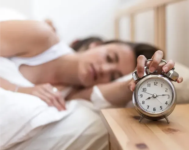 Een vrouw in bed probeert een wekker op het nachtkastje uit te zetten.