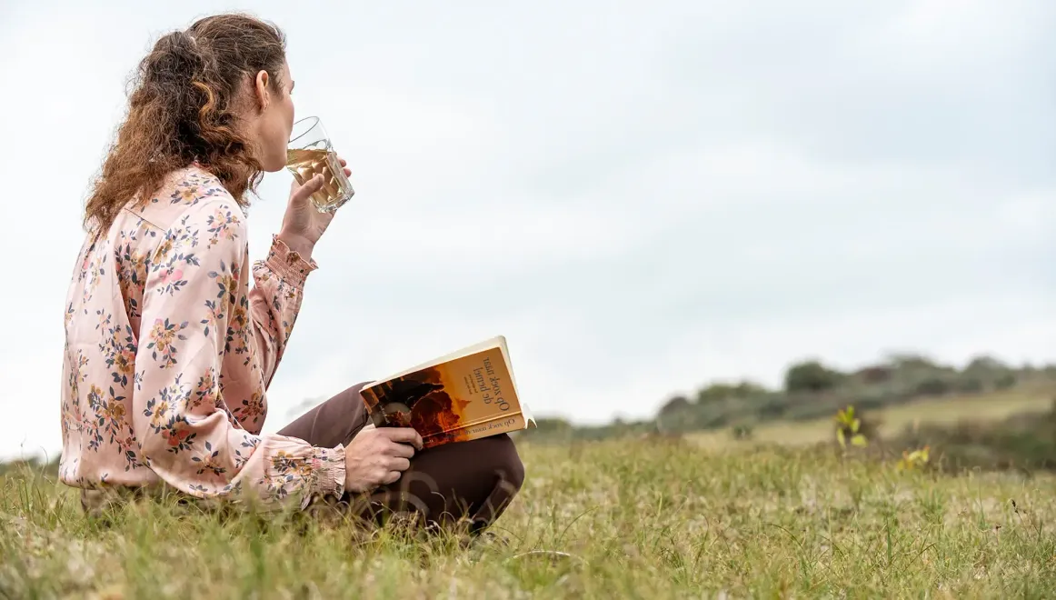 Annemarie drinkt uit een glas, zittend op een met gras begroeide heuvel naast een honingraatstructuur. Bewolkte lucht.