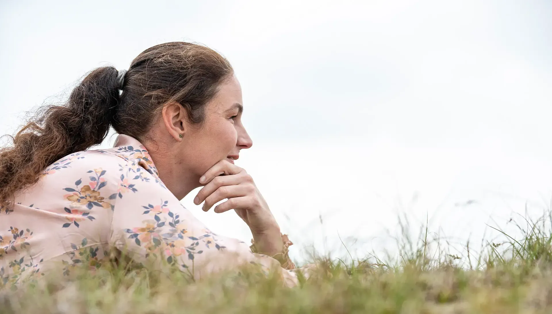 Annemarie met krullend bruin haar, kin op haar hand rustend, liggend in het gras, kijkend naar de lucht.