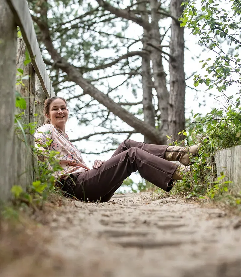 Lachende Annemarie, zittend op een houten pad, leunend tegen een leuning. Bruine broek, bloemetjesblouse, natuurlijke omgeving.