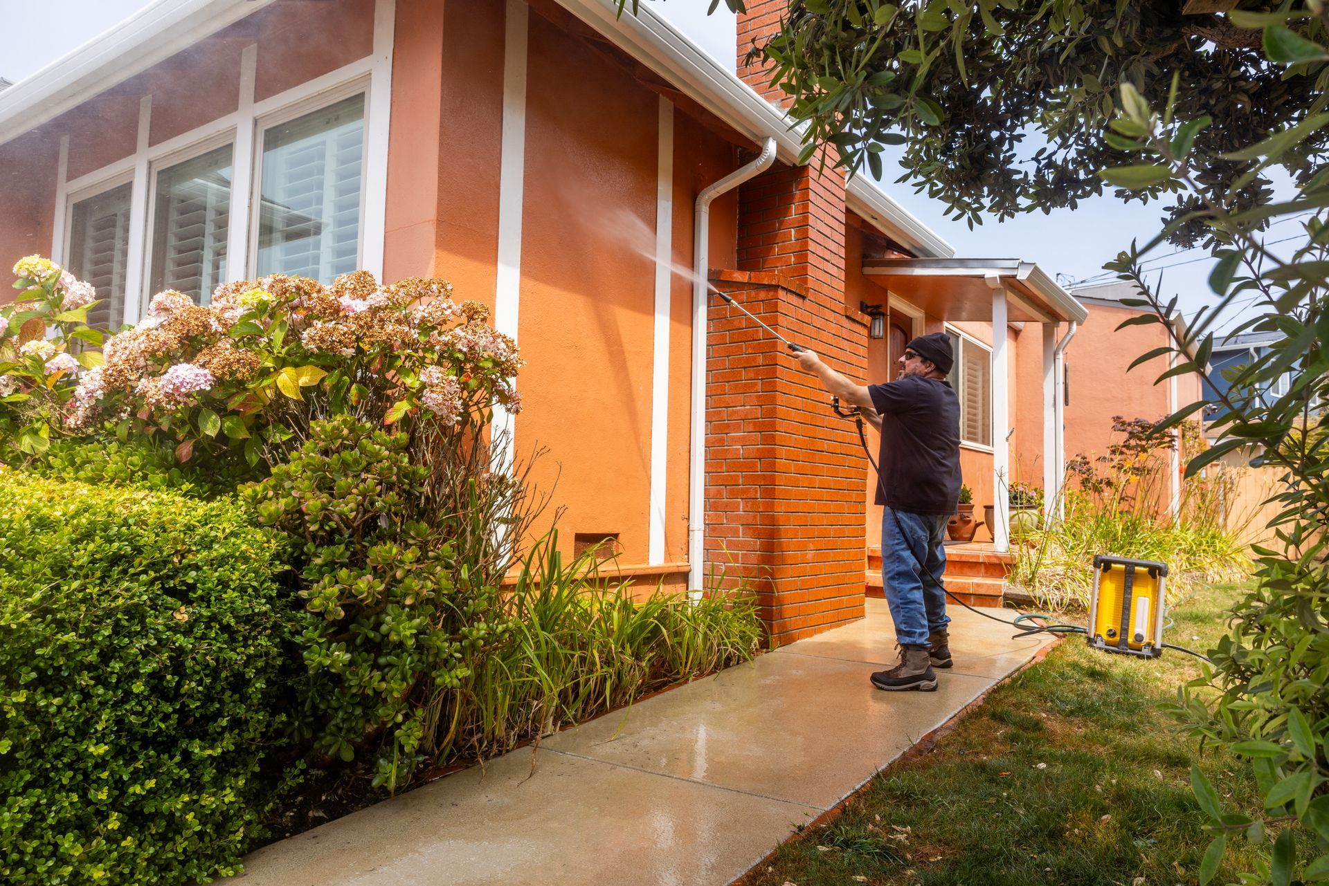 A man is using a pressure washer to clean the sidewalk in front of a brick house.
