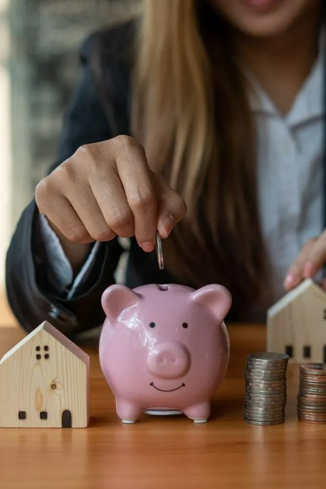 Woman Putting Coin in Pink Piggy Bank with House Models — All Finanz & Insurance Solutions Mackay in North Mackay, QLD