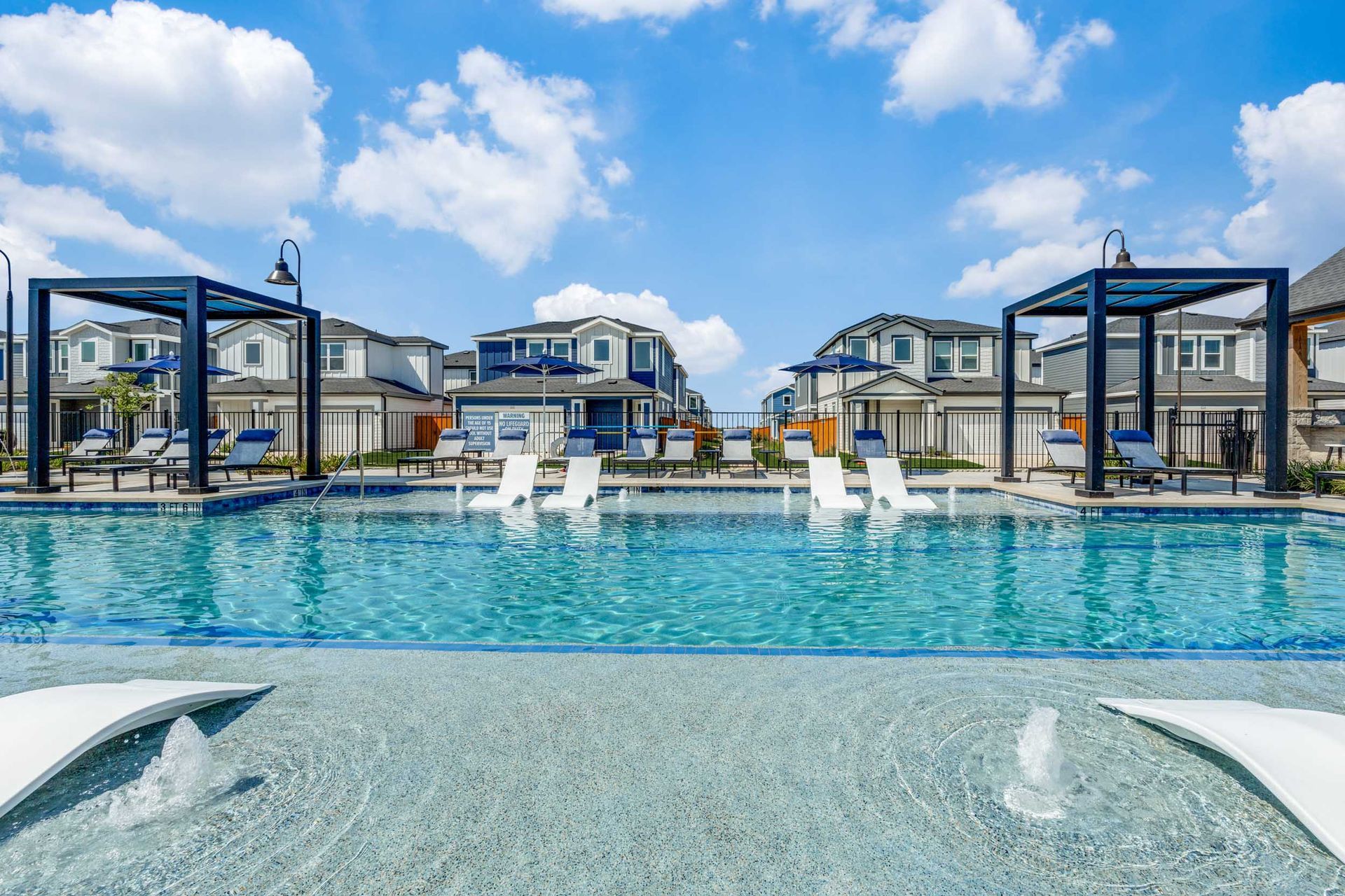 Swimming pool with lounge chairs, houses in the background under a bright blue sky.