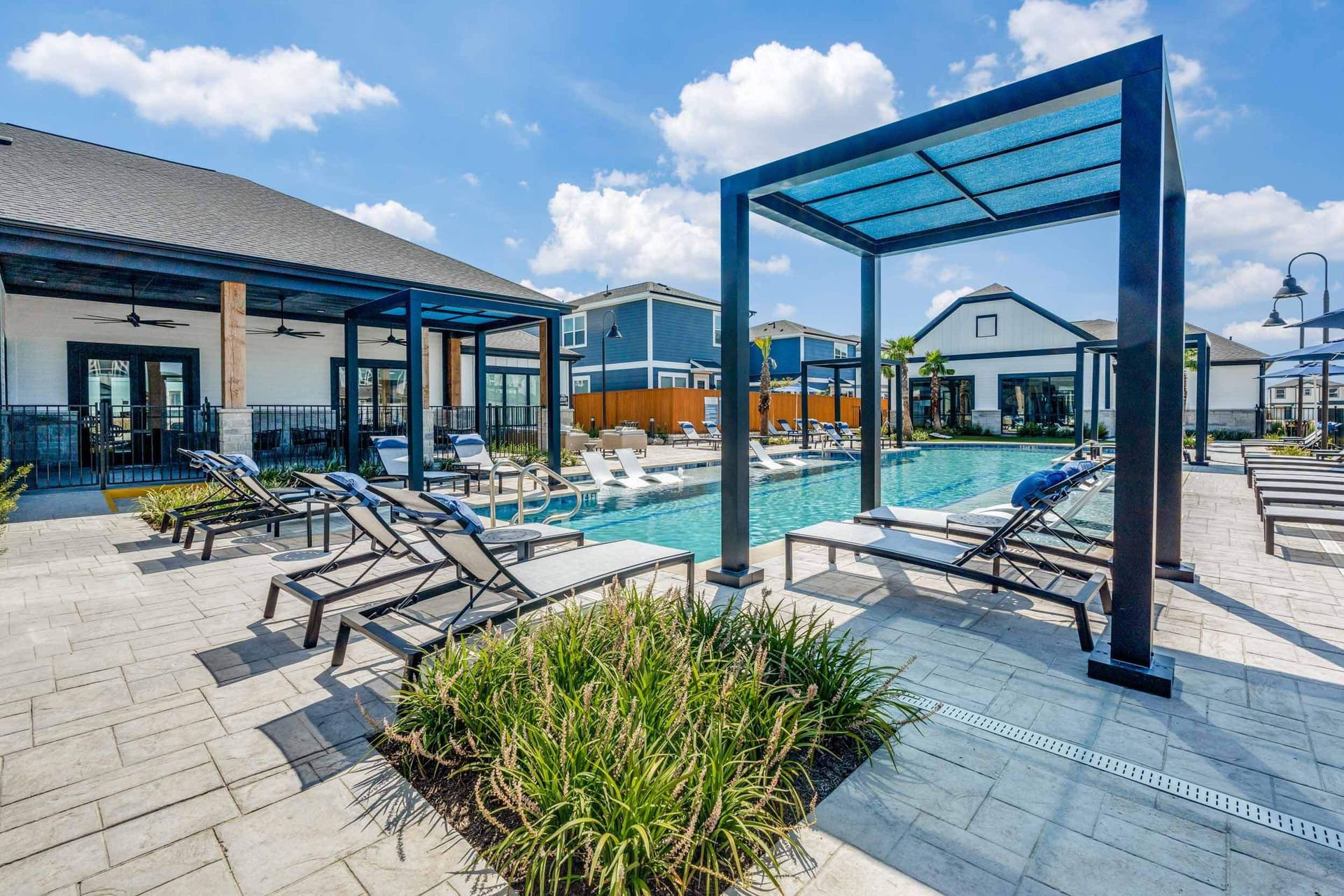 Poolside lounge area with a pergola, chaise lounges, and a building. Blue sky.
