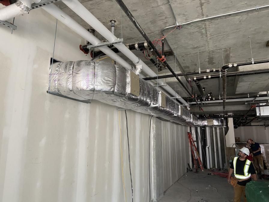 Construction site: ductwork wrapped in silver insulation runs along the wall and ceiling. Workers are present.