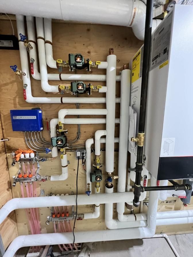 Plumbing system with white insulated pipes, valves, pumps, and controls mounted on a wooden wall.