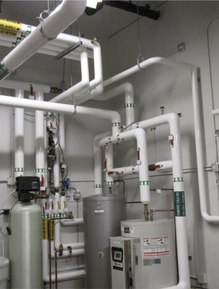 Pipes and water heaters in a utility room. White pipes, silver and gray tanks, and various gauges are visible.