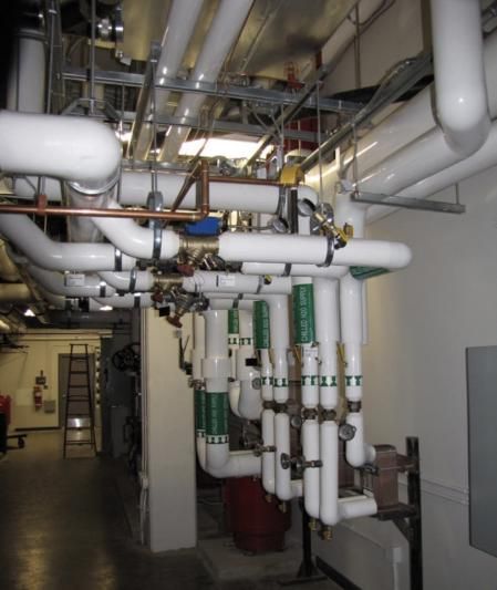 Pipes and valves in a mechanical room. White pipes, green valve tags, and a gray metal ceiling.