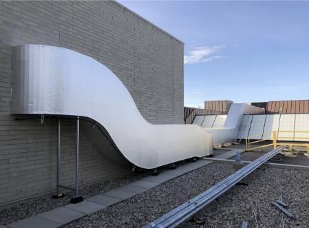 Aluminum HVAC ductwork curves along a brick wall and across a rooftop under a blue sky.