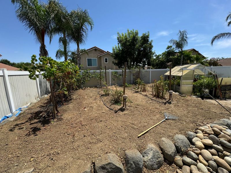 A backyard with a fence , trees , rocks and a house in the background.