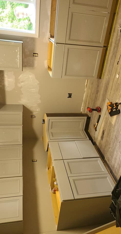 Kitchen cabinets being installed in a room with unfinished walls and a window; light gray cabinets.