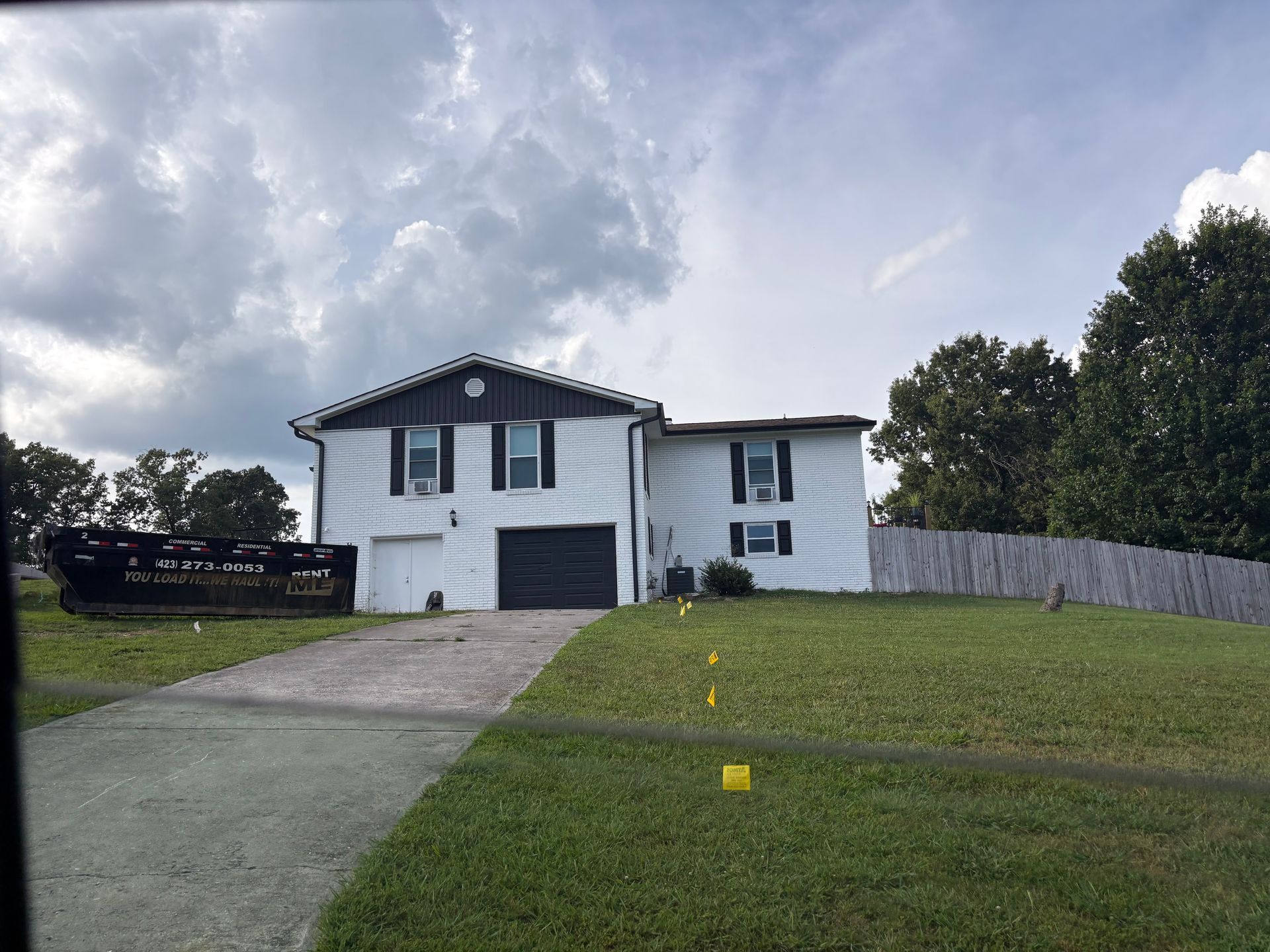 White two-story house with black shutters and garage. A driveway leads to the house; green lawn with cloudy sky.