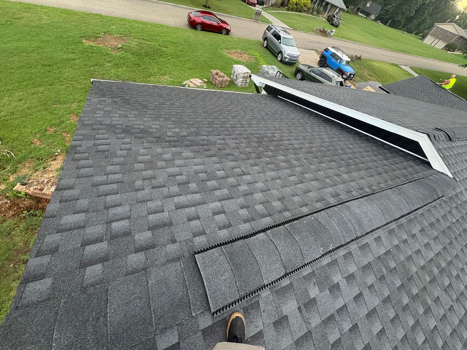 View of a dark gray shingle roof with unfinished sections and a vent. Cars and green yard in background.