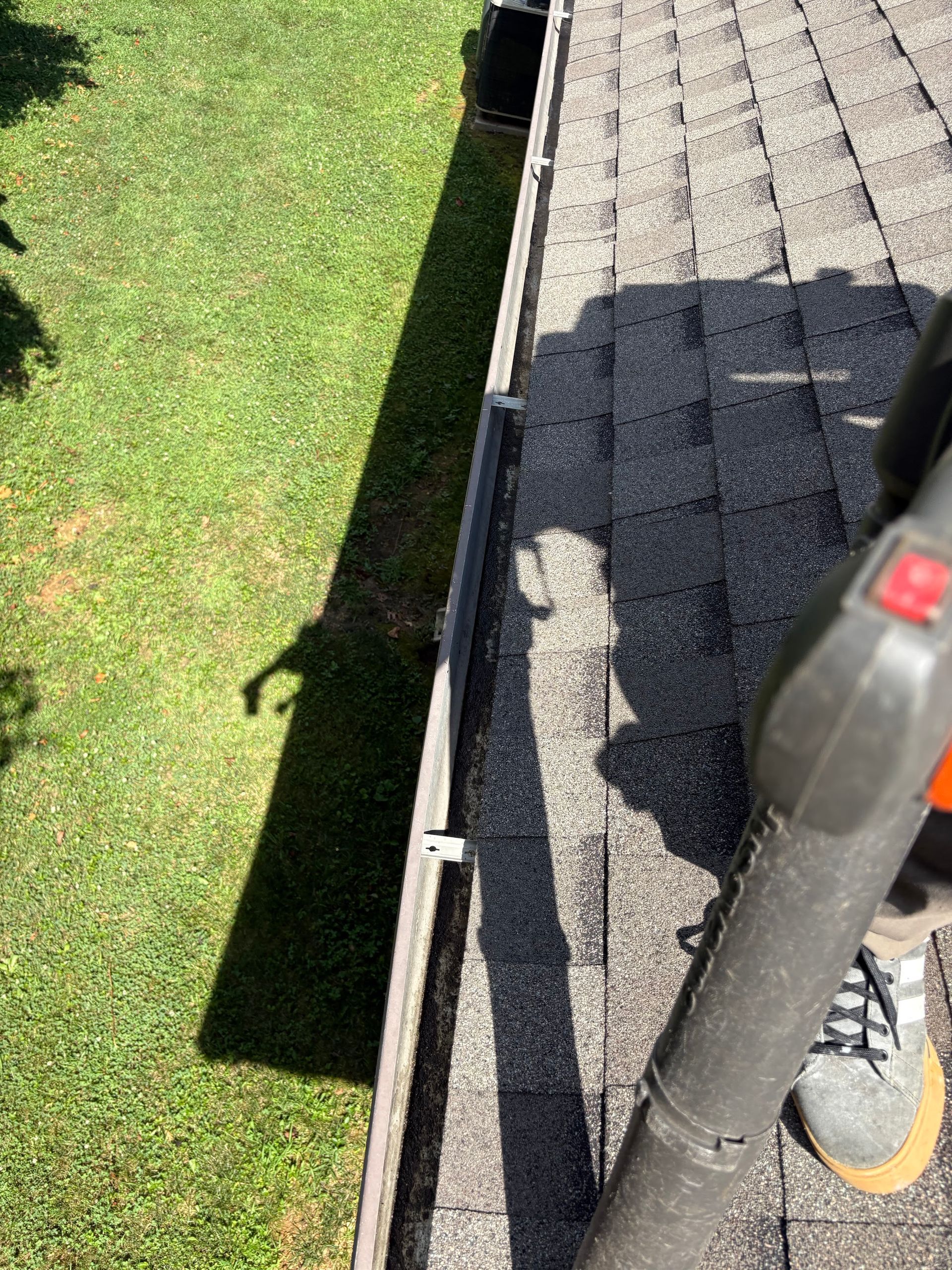Person using a leaf blower to clean a gutter on a roof. Shadow of the person on the grass.