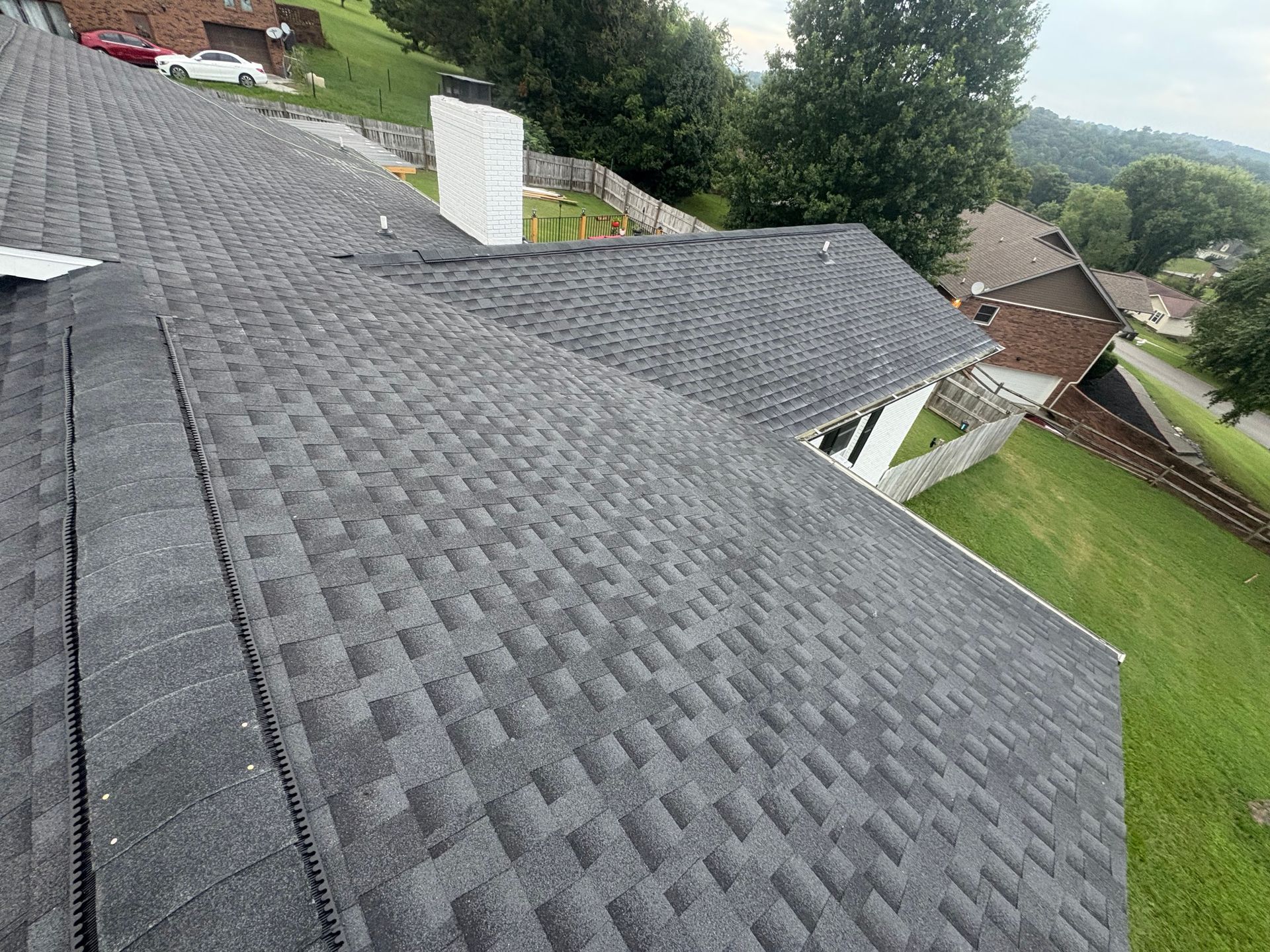 Dark asphalt shingle roof of a house with a white chimney, green lawn, and surrounding trees.