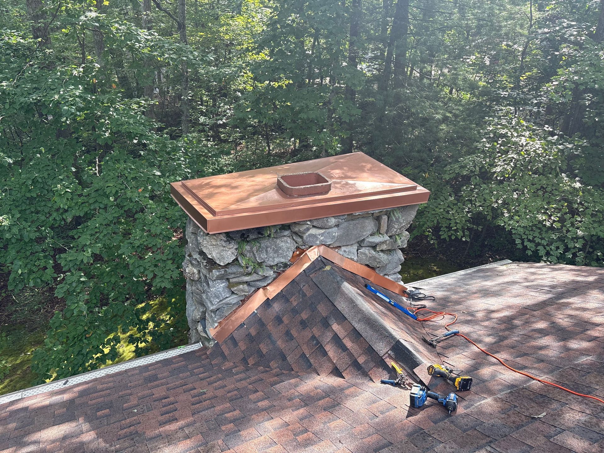 Stone chimney with copper cap and roof shingles, surrounded by green trees.