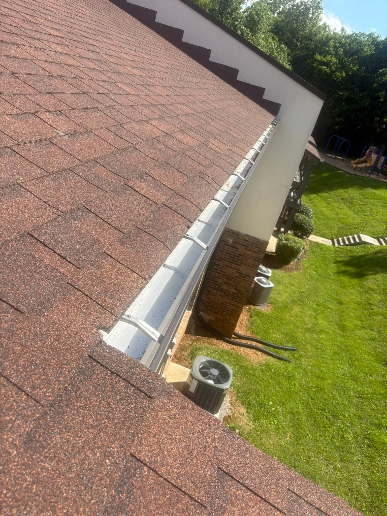 Brown shingled roof with a white gutter and brick chimney. Green grass and trees in the background.