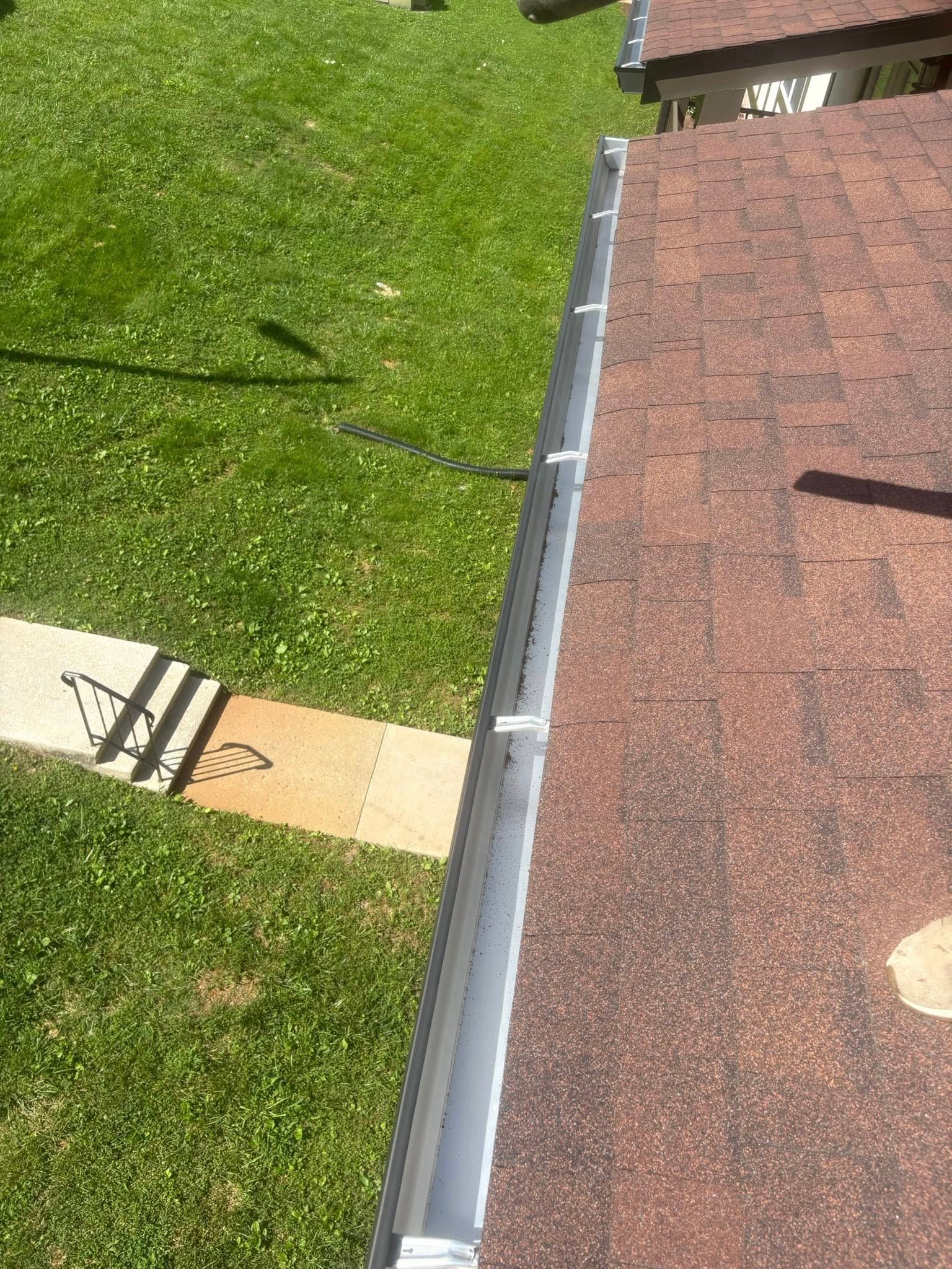 Gutter along a brown shingle roof, with green grass and a sidewalk in the background on a sunny day.