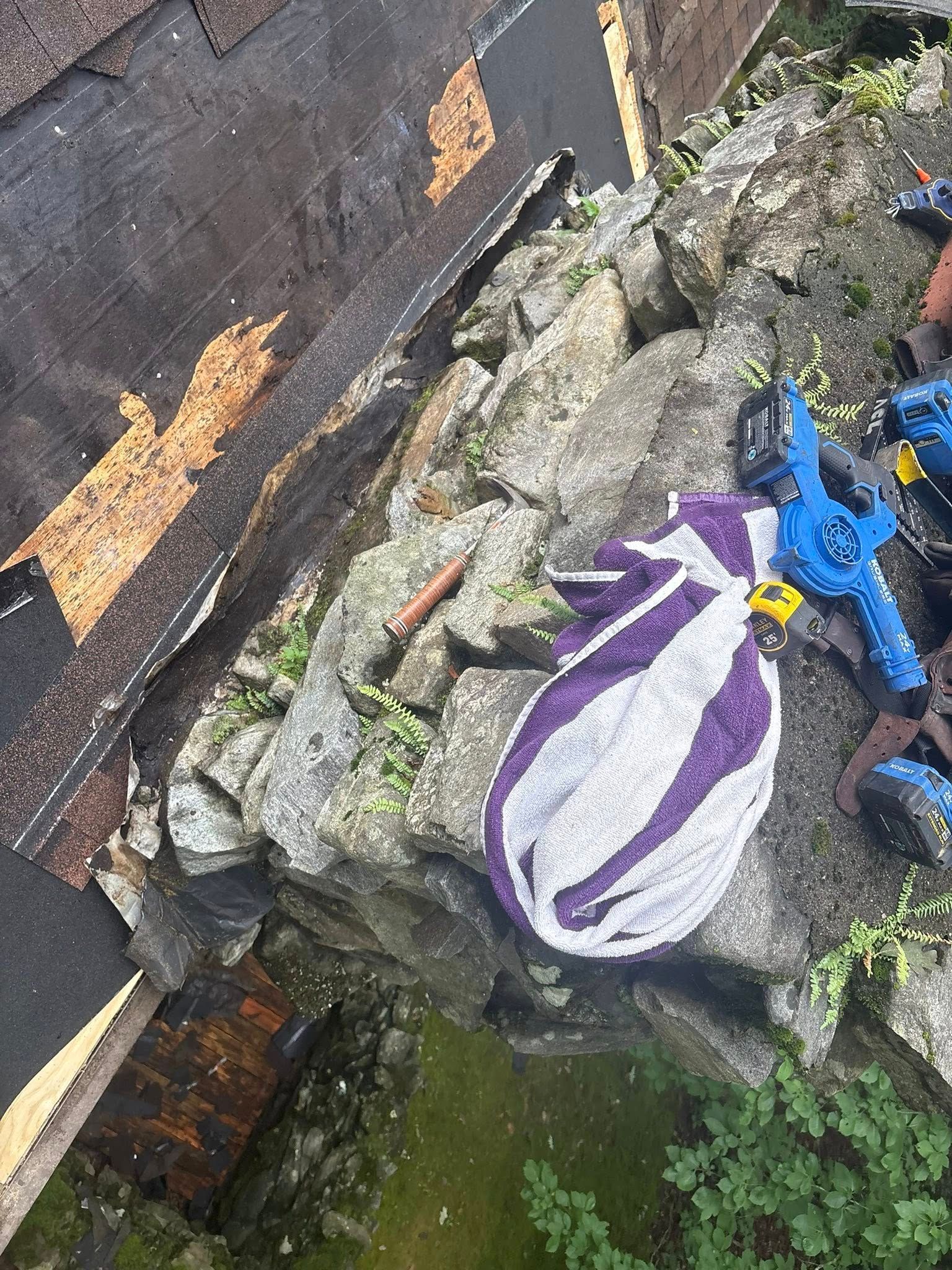 Rooftop with damaged shingles next to a rock wall; purple and white bag sits on the rocks, possibly trash.