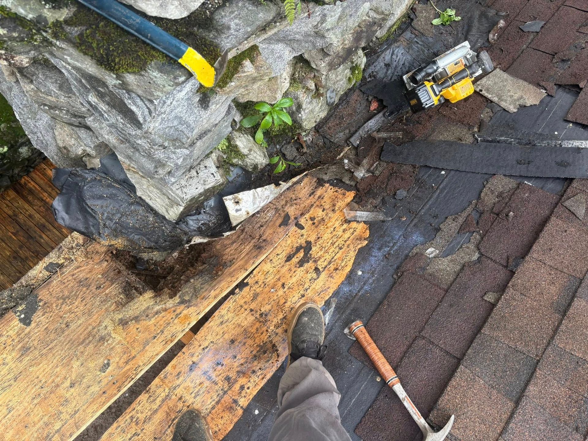 Roof repair scene: worker on rooftop, removing old shingles and wood near a stone wall, using tools.