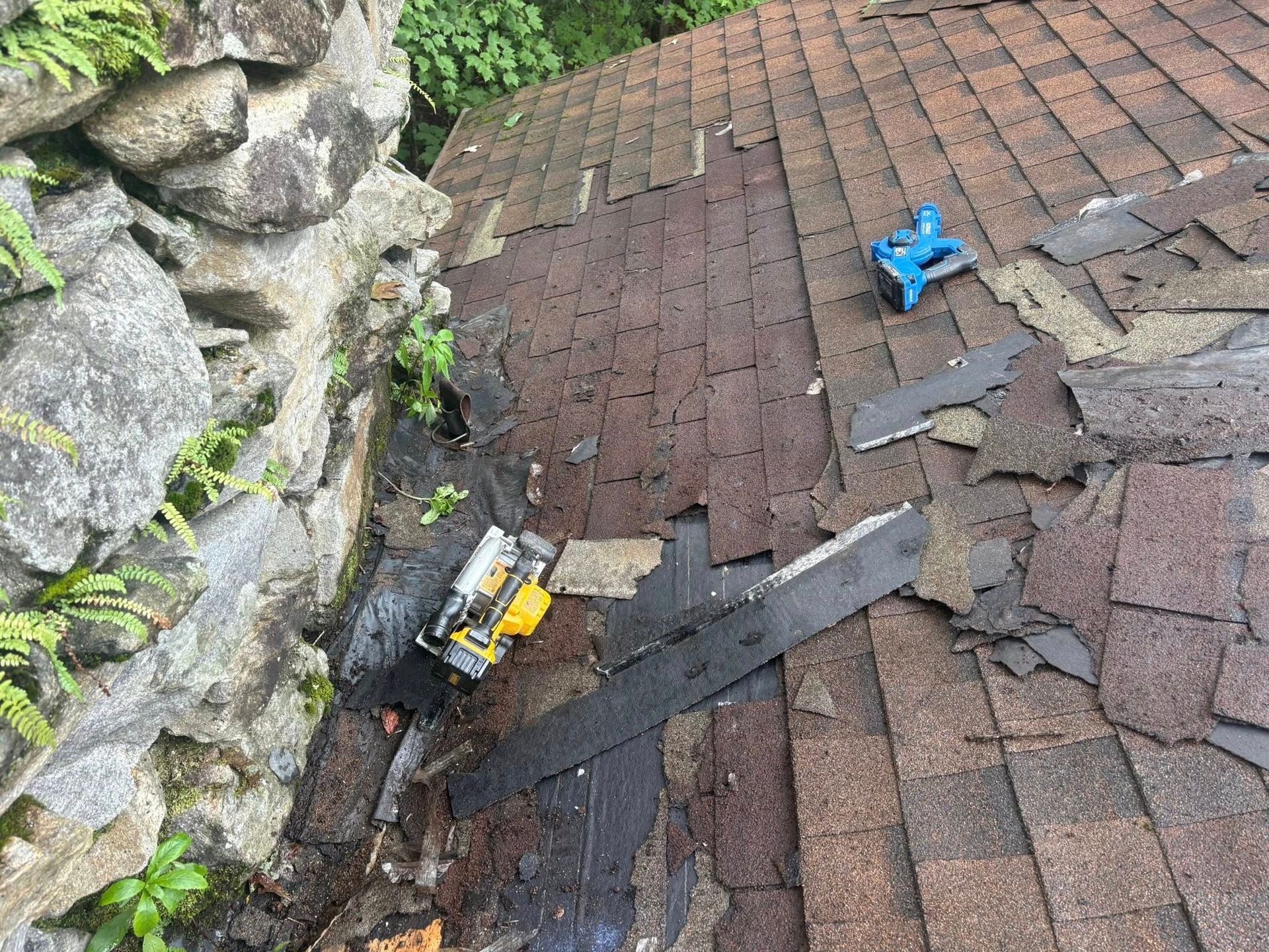 Roof with missing shingles and tools. A stone wall is on the left. Yellow saw, blue tool, and roof damage are visible.
