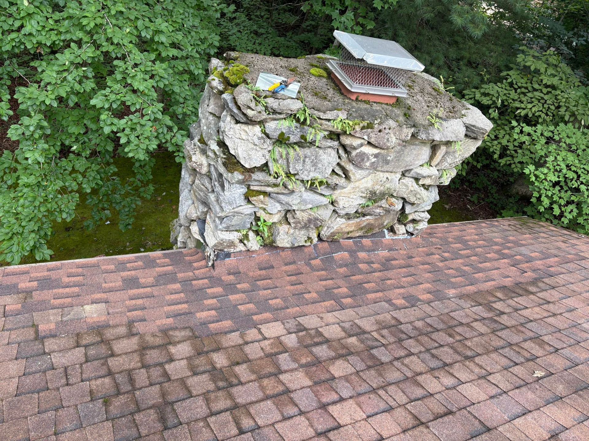 Stone chimney on a roof with shingle tiles, surrounded by green foliage.