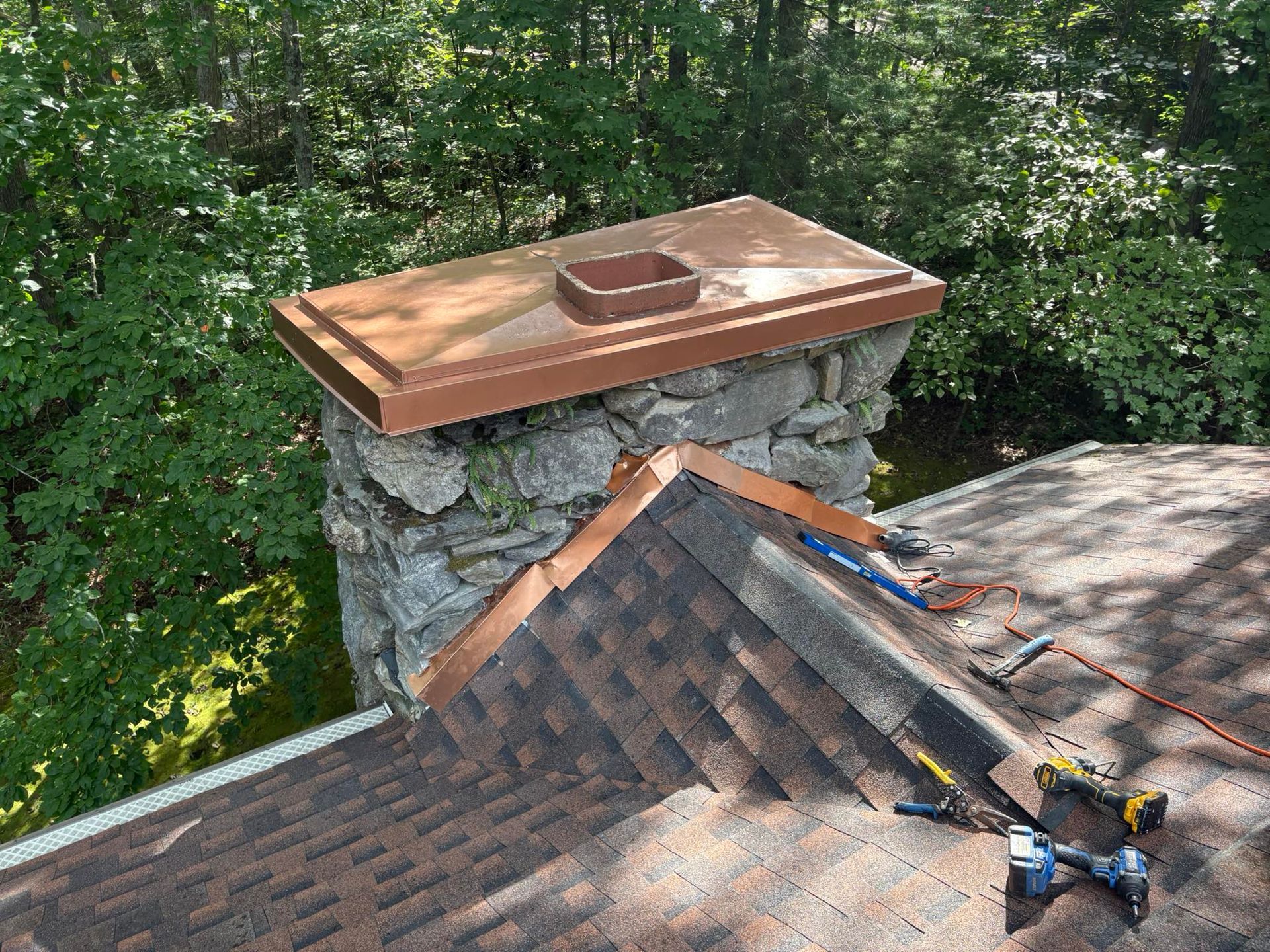 Chimney with copper cap on a shingled roof, tools nearby, surrounded by trees.