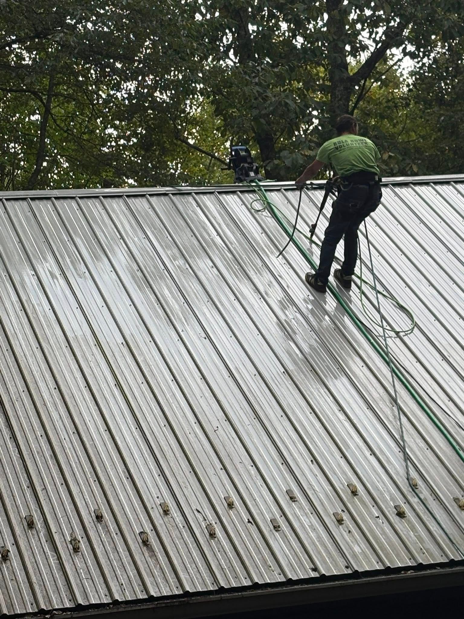 Two workers on a metal roof cleaning with hoses, secured by ropes, surrounded by trees.