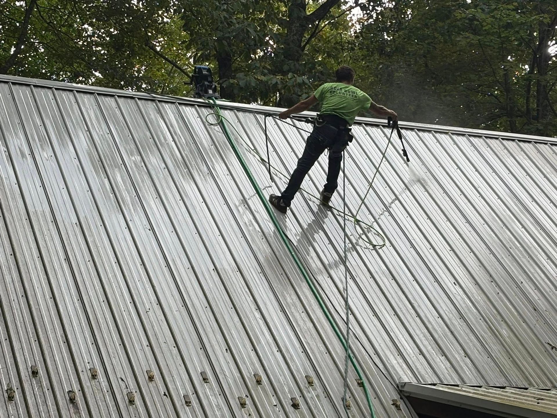Person spraying down a metal roof, wearing safety gear. Outdoors, daylight.