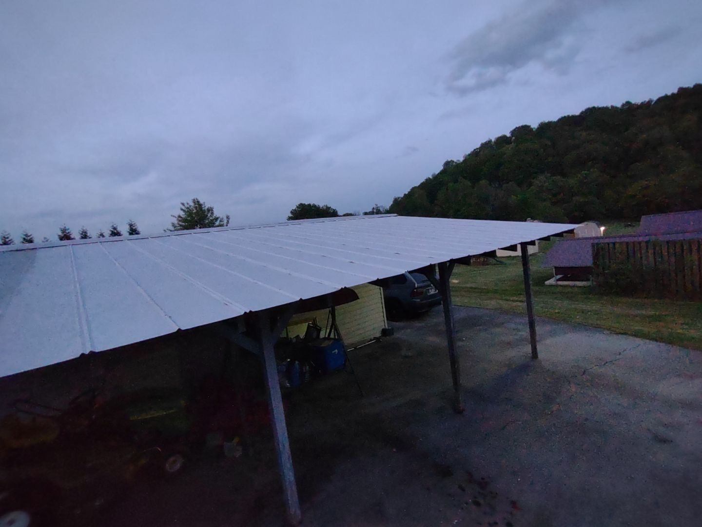 Metal roof carport, parked car, cloudy sky, hill in background.