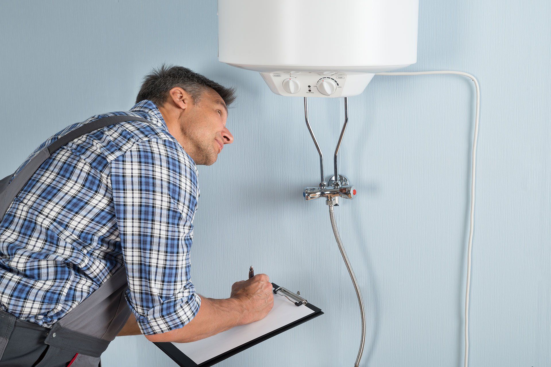 A plumber in blue plaid shirt inspects a water heater, taking notes on a clipboard.