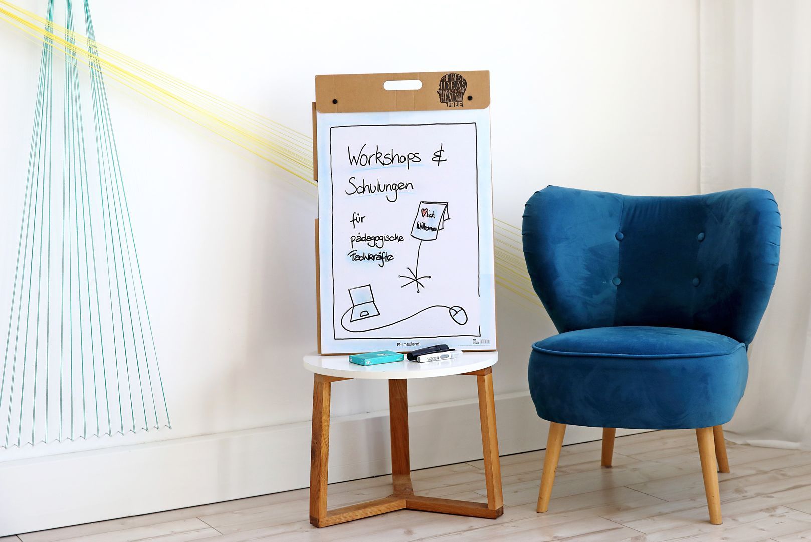 Der blaue Sessel steht neben einem Tischchen. Darauf siehst du eine Flipchart mit einem Willkommensplakat. 