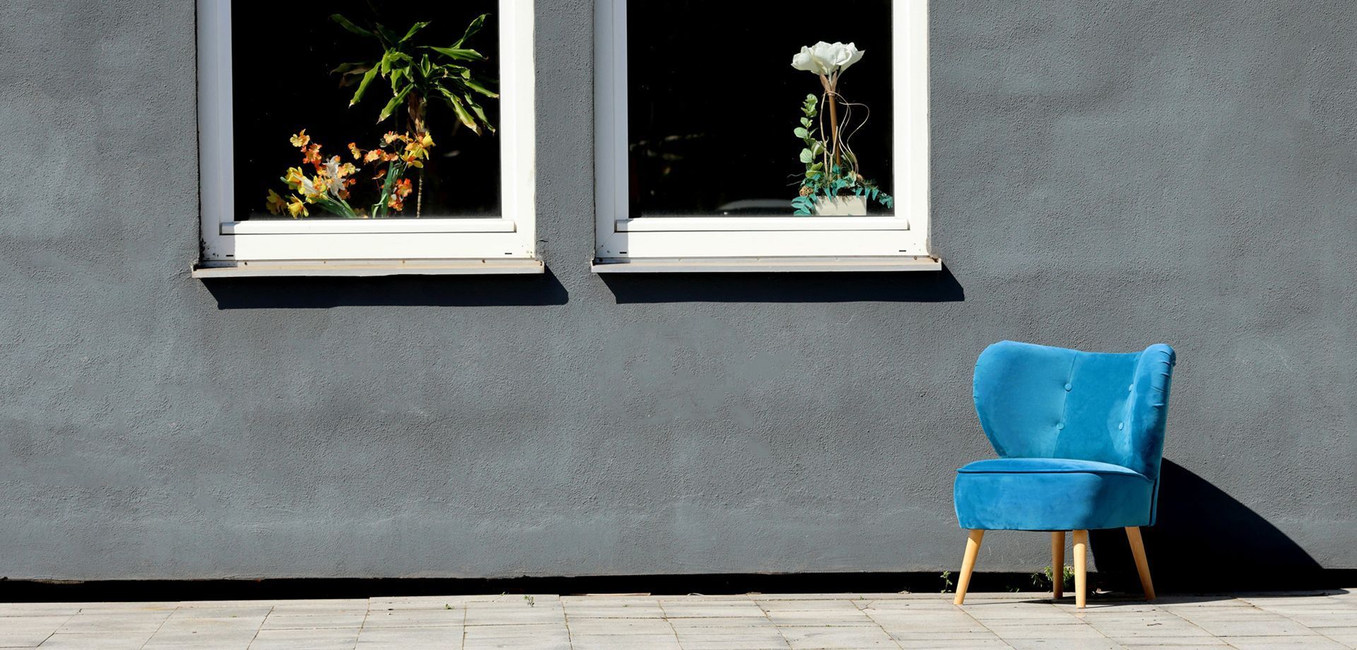 Der blaue Sessel steht vor einer Hausfront. In den beiden Fenstern siehst du Blumen. 