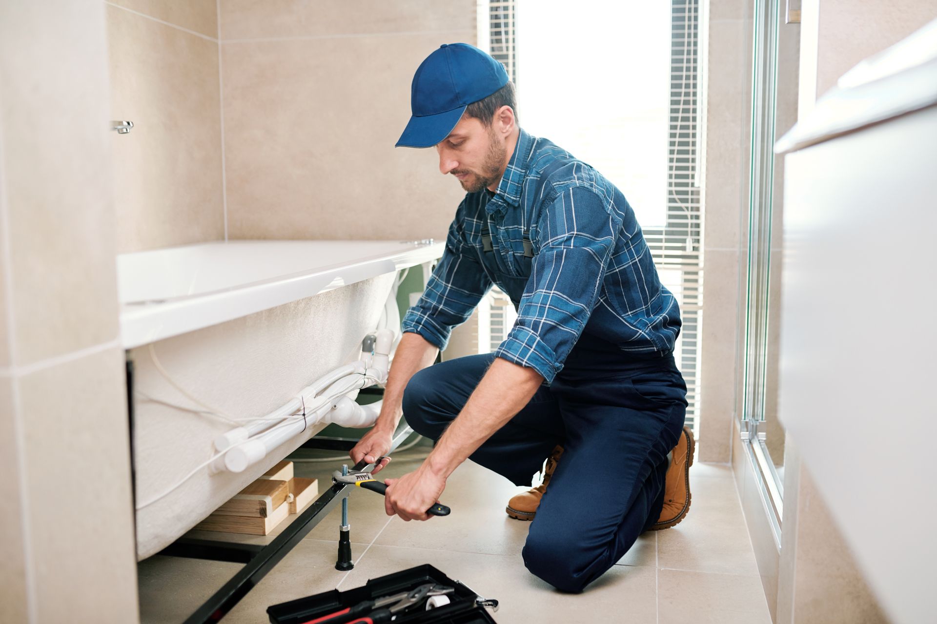Plumber in blue uniform kneels, working on pipes under a bathtub in a bathroom.