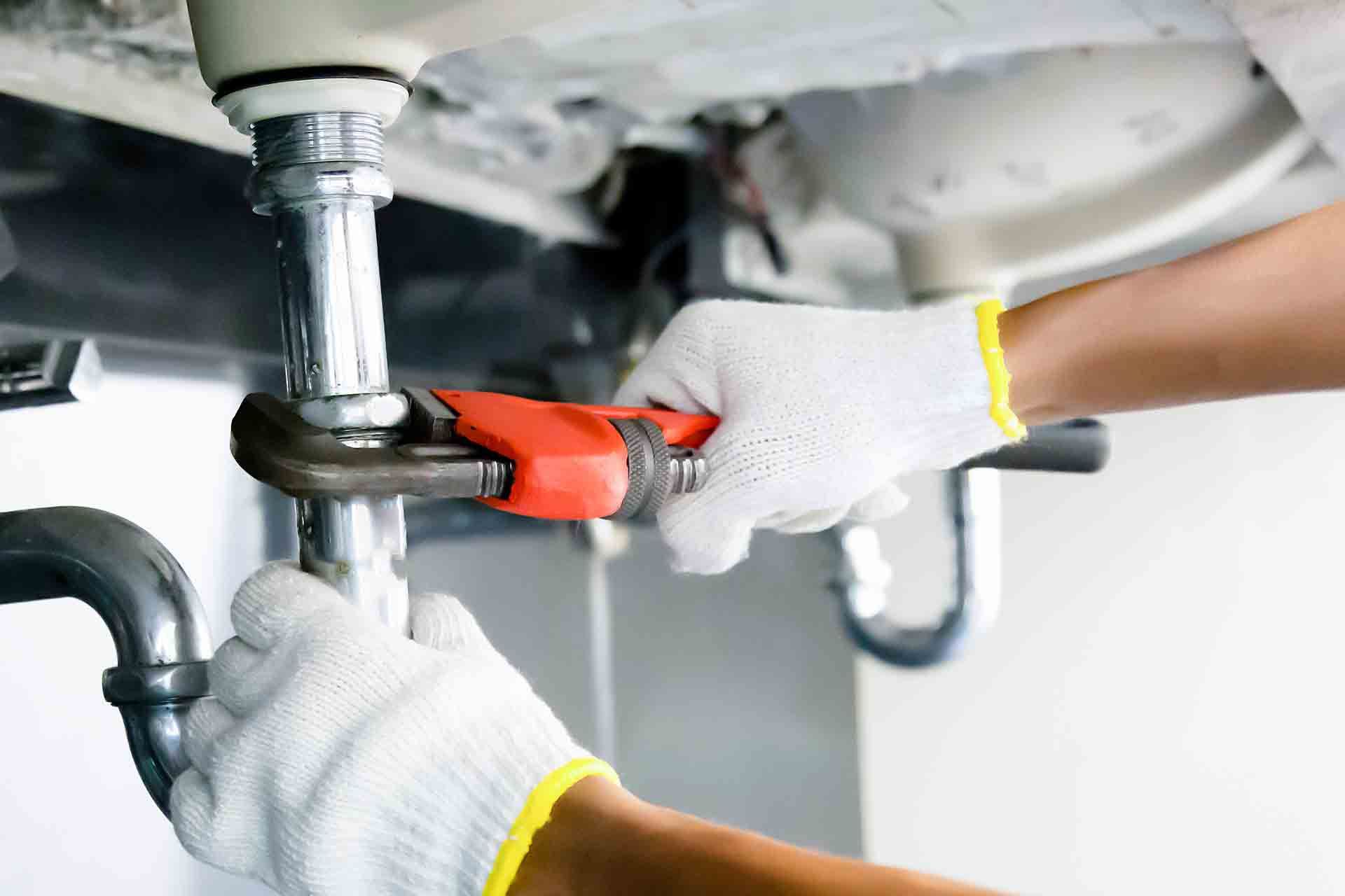 Plumber with gloves using wrench to tighten pipes under a sink.