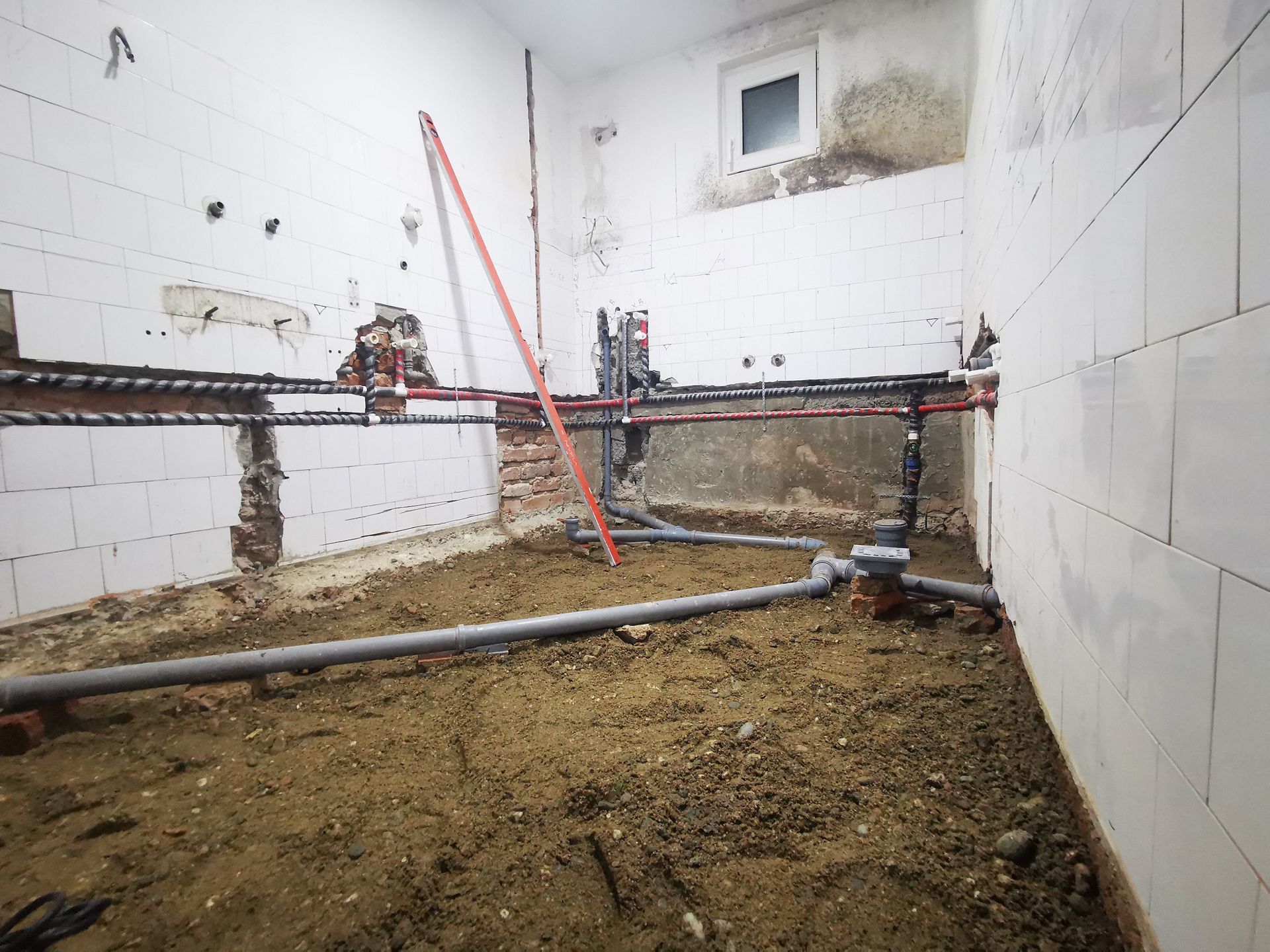 Bathroom renovation: exposed pipes, dirt floor, partially tiled walls, level in place.