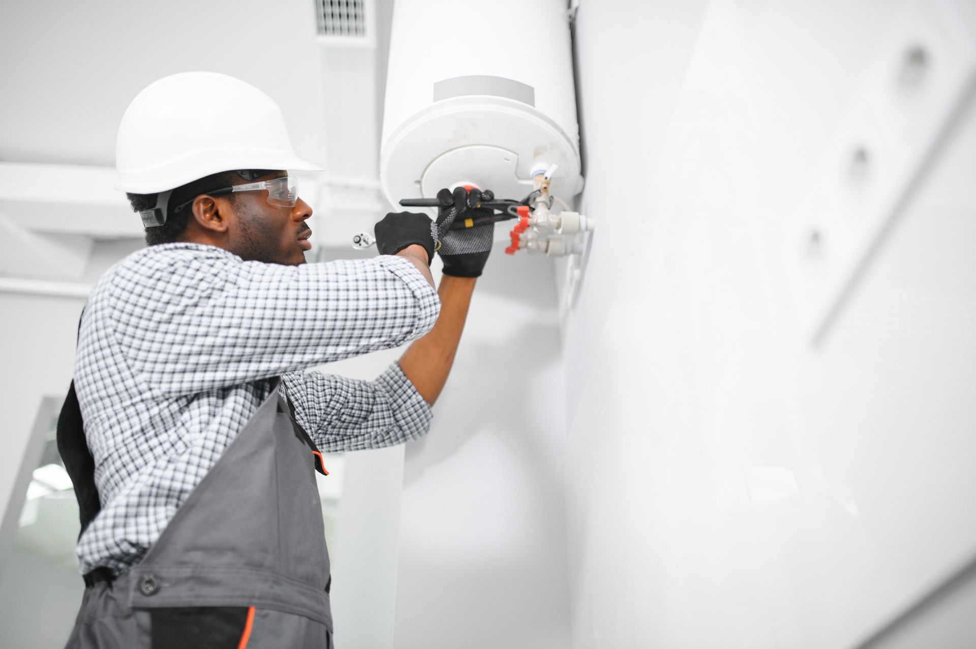 Electrician in a white hard hat and goggles working on a water heater, white wall.