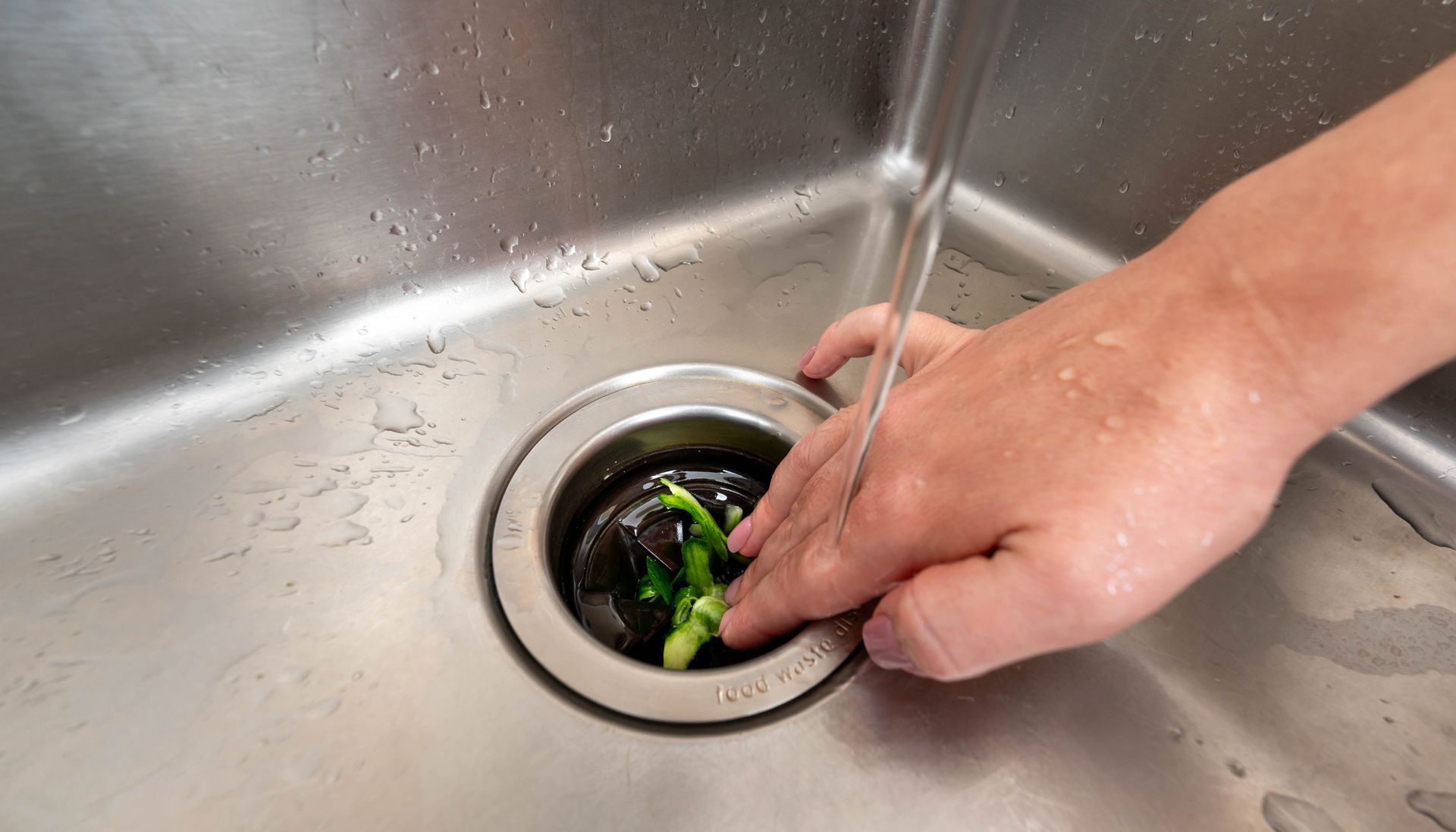 Hand washing green vegetable scraps down a kitchen sink drain with running water.