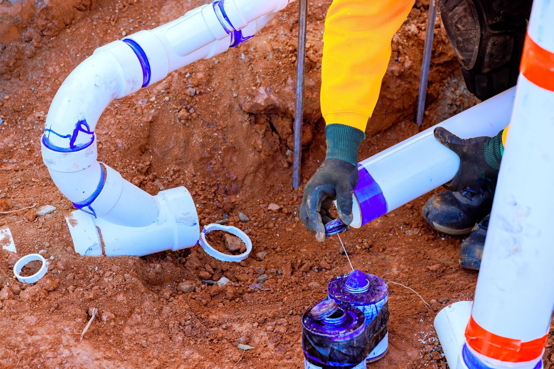 Plumber connecting white PVC pipes in a dirt trench, using purple primer.