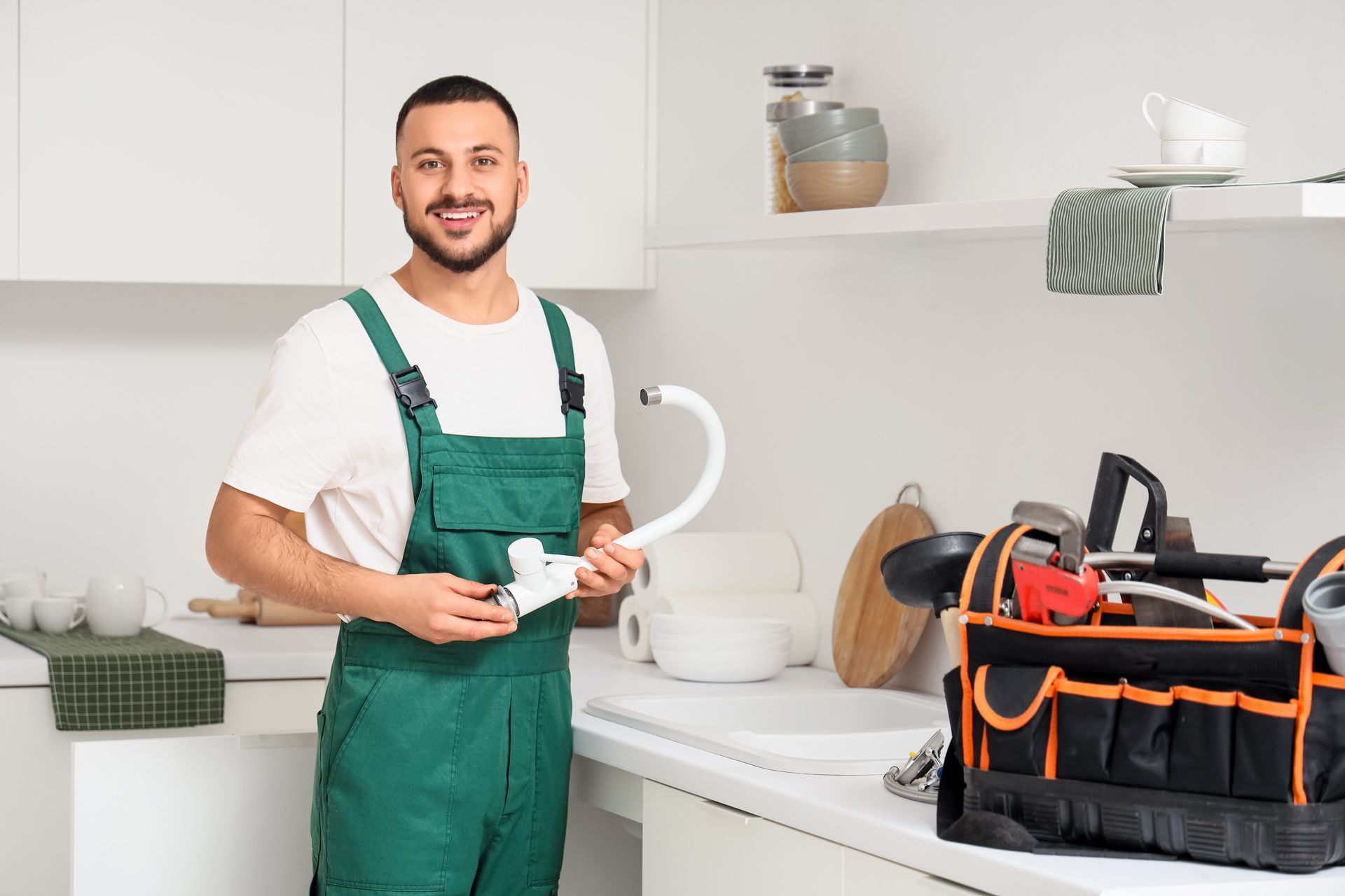 Plumber in blue shirt and overalls works on a kitchen faucet in a kitchen.