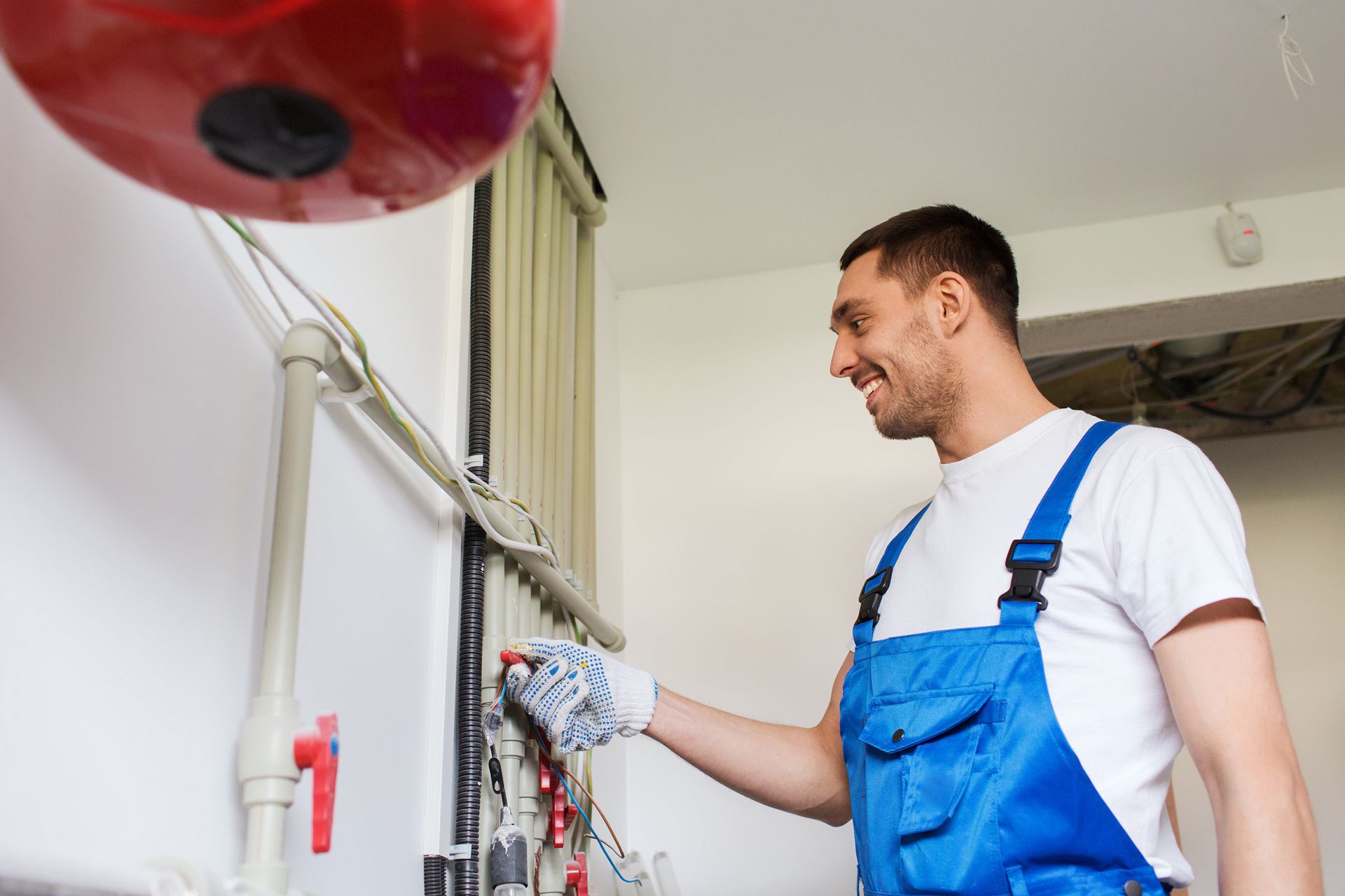 Plumber in blue overalls, smiling, working on pipes near a red component.