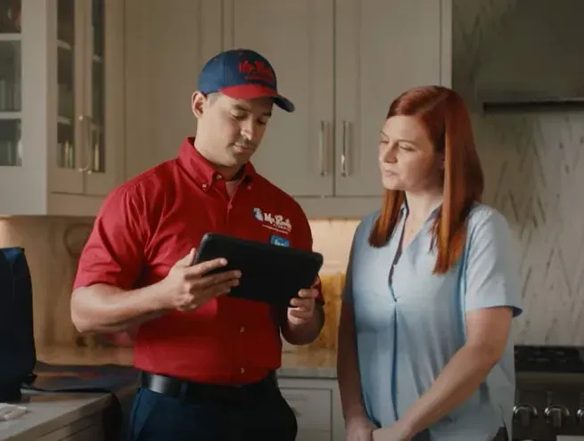 A professional wearing a red uniform and cap shows a tablet screen to a person while standing in a home.