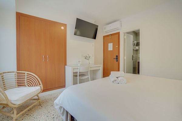 Habitación de hotel pequeña con cama, armario, televisor de pared, escritorio y silla en tonos blancos y de madera.