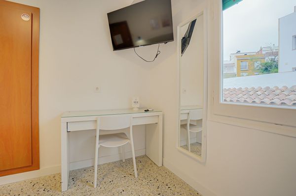 Habitación minimalista blanca con escritorio, silla, televisor montado en la pared, espejo y vista a la ventana.